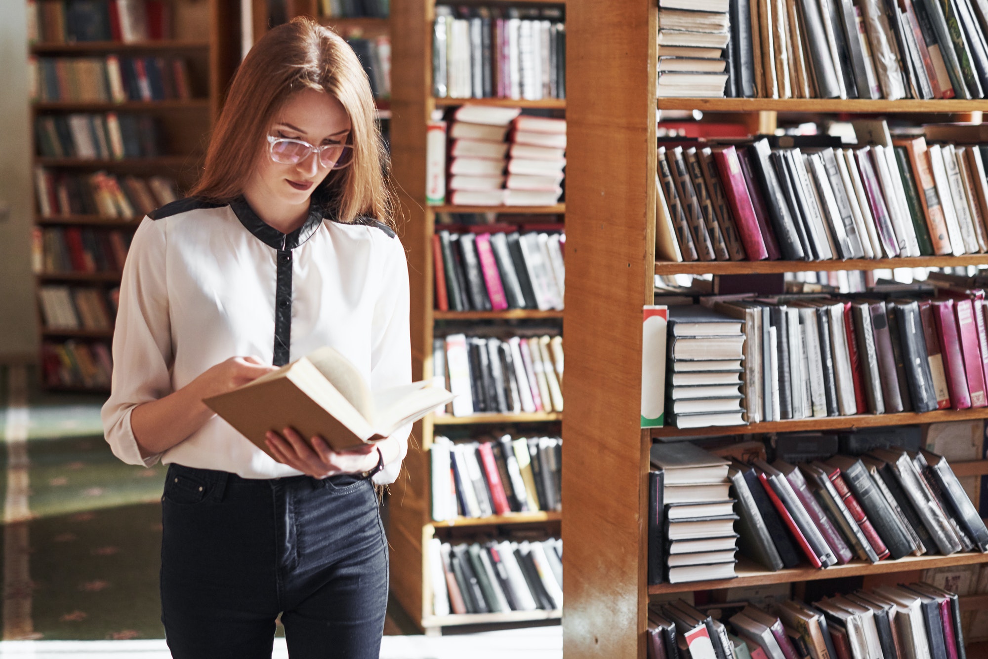Young attractive student librarian reading a book between library bookshelves