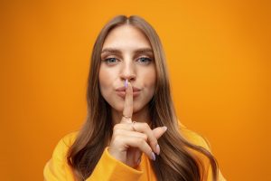 Pretty young woman holding finger on lips and showing silence sign on yellow background