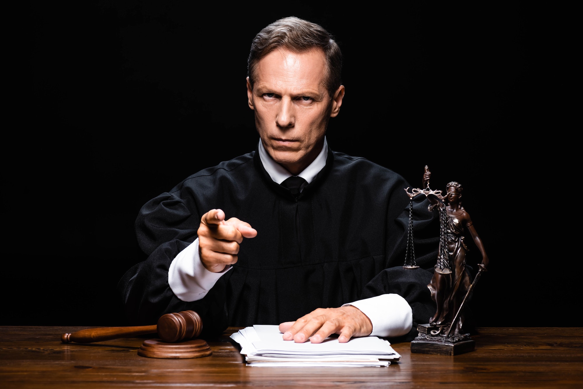 judge in judicial robe sitting at table and pointing with finger isolated on black