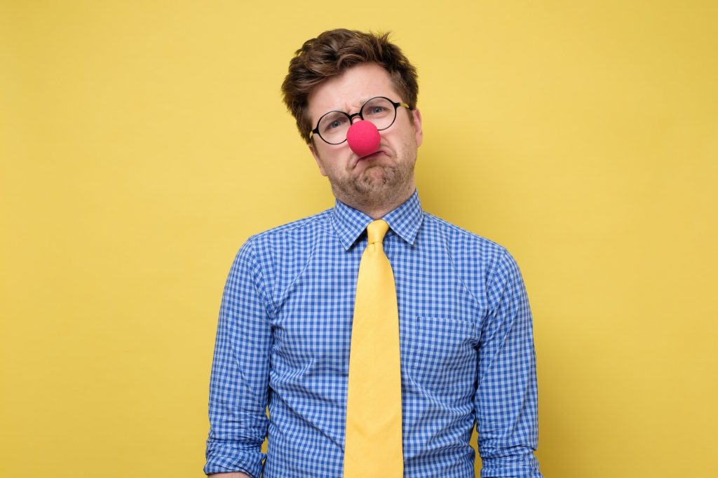 Sad caucasian man wearing clown red nose on yellow wall.