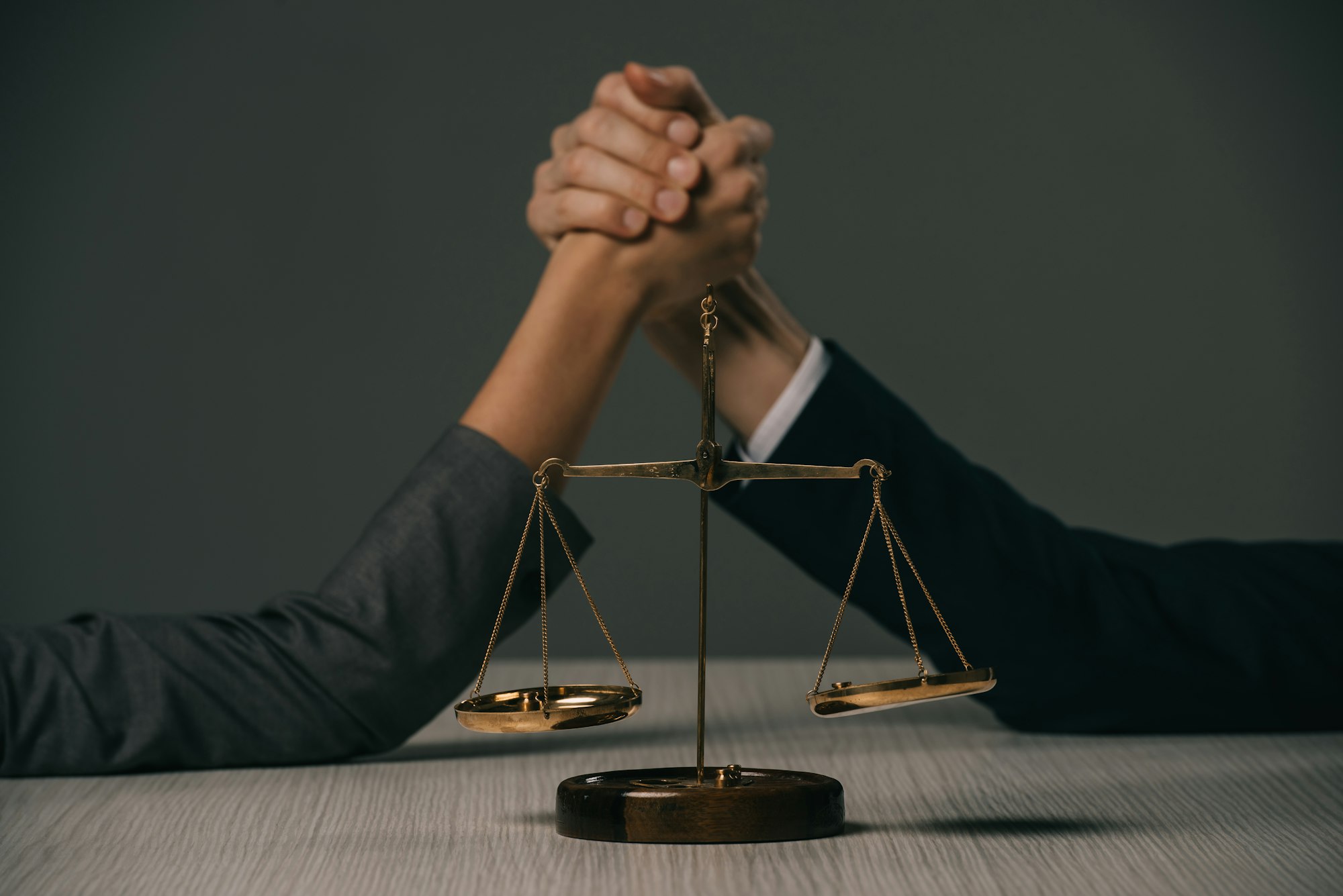 cropped view of businesspeople arm wretsling on wooden table with scales of justice on grey